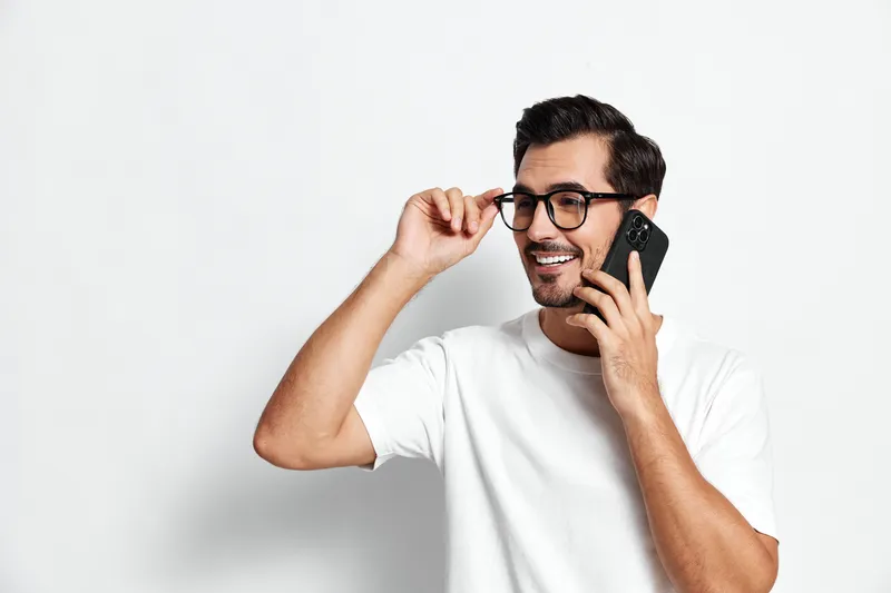 Smiling man wearing glasses talking on mobile phone with cheerful expression against plain light background. People lifestyle concept