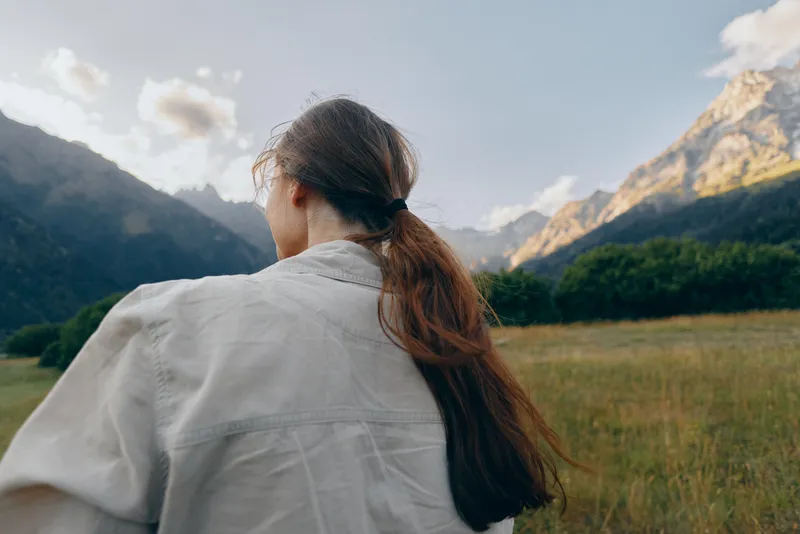 Young woman with long brown hair stands in a wide open field facing distant mountains under a partly cloudy sky, enjoying a peaceful moment in nature.