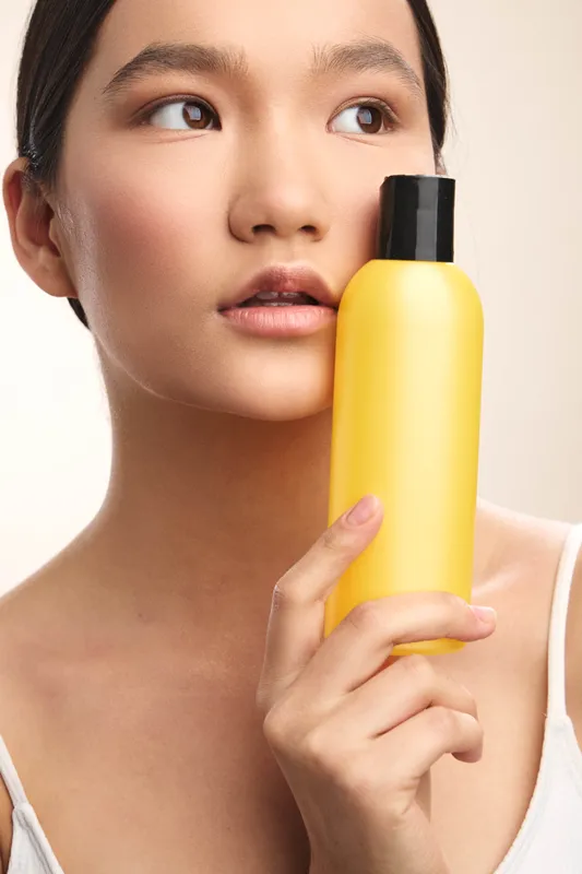 asian woman holds a yellow bottle near her face in a close up portrait, conveying beauty care focus and calm confidence in a studio setting.