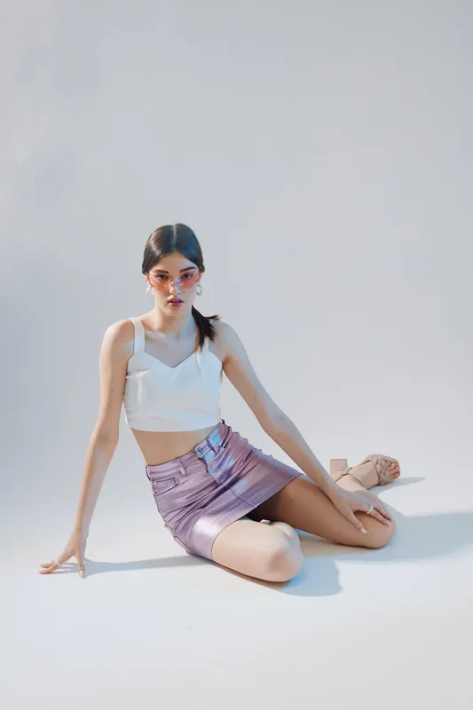 Young woman in fashion outfit poses sitting on the floor in bright studio. She wears a white top and purple skirt with stylish sandals. Concept of beauty and fashion.