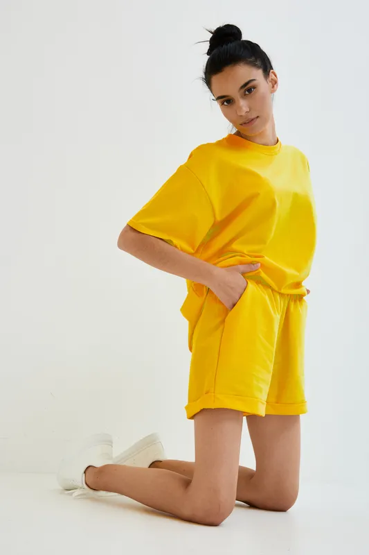 Young woman in bright yellow casual outfit kneeling against white background, showcasing sporty summer style with minimalist composition and relaxed mood.