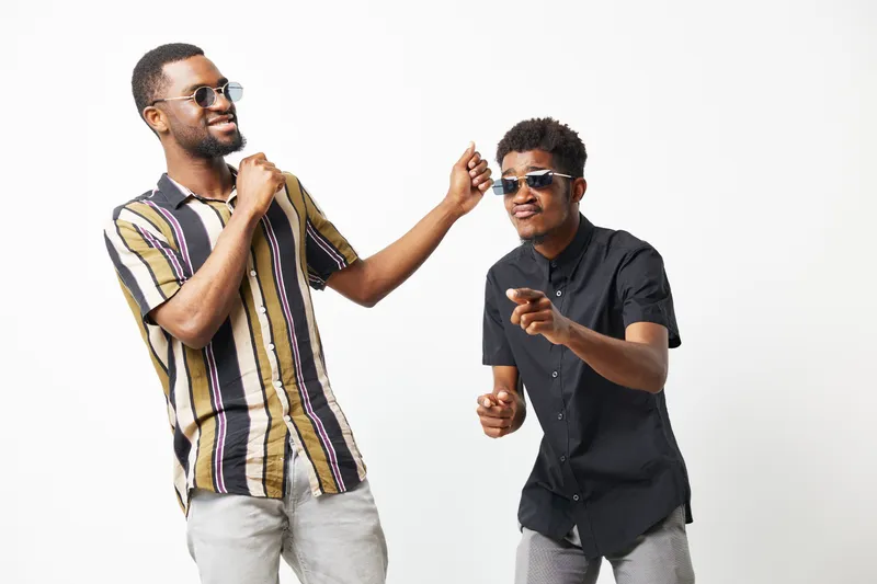 Two joyful black men dancing with sunglasses, showcasing friendship and fun. Their playful energy highlights modern fashion in a bright, minimalist setting.