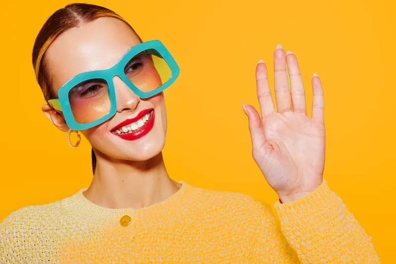 Smiling woman with red lipstick wearing large blue sunglasses and yellow sweater waves hand on bright orange background. Concept of fashion, summer, happiness, and vibrant style.