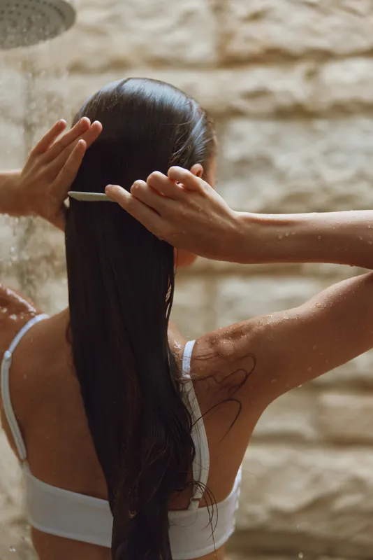 Woman combing long wet hair under warm outdoor shower, natural light, bare skin, peaceful moment, summer wellness and self-care routine.