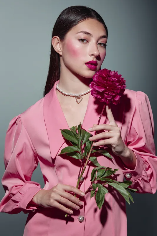 Elegant young woman wearing pink fashionable jacket holds bright vibrant pink peony flower close. Makeup is bold with dark lips and cheeks. Studio portrait with creative beauty concept.