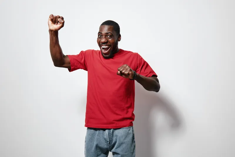 African American man with dark skin wearing red t shirt and jeans smiling and celebrating with raised fist on white background. Happy, excited, casual and energetic male portrait.