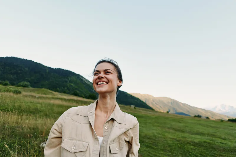 Smiling woman in casual jacket standing outdoors in a green field with mountains in the background. Concept of lifestyle, nature, happiness, and freedom in soft natural light.
