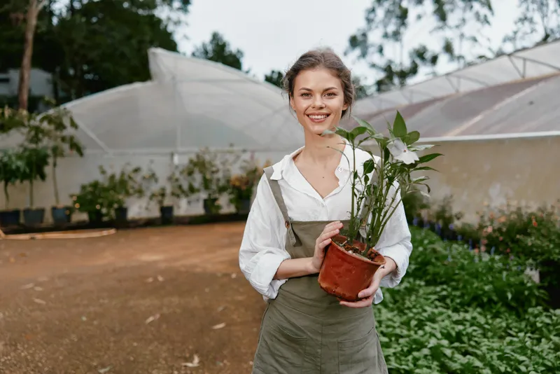 Young woman smiling while holding a potted plant in a greenhouse, showcasing gardening passion and connection with nature, ideal for eco friendly themes