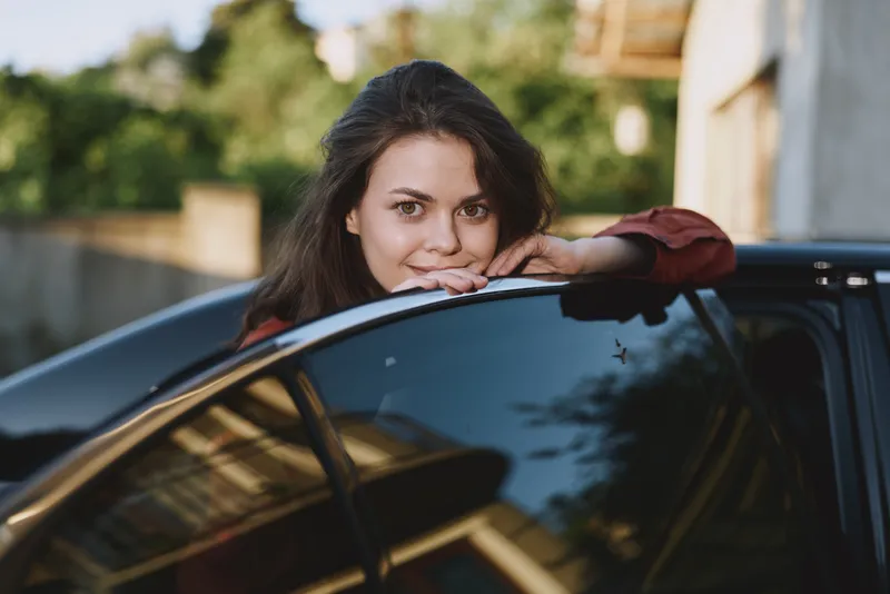 Smiling woman leaning on a car window outdoors, capturing a casual lifestyle moment, relaxed confidence, and everyday urban energy. Soft lighting adds mood today