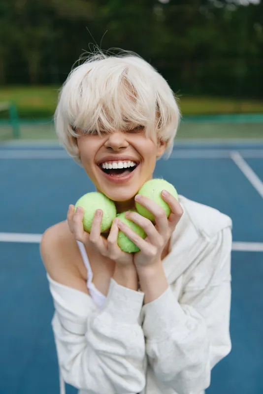 Joyful blonde woman holding green tennis balls, smiling widely in a sporty outfit against a blue tennis court background, reflecting energy and enthusiasm for sports
