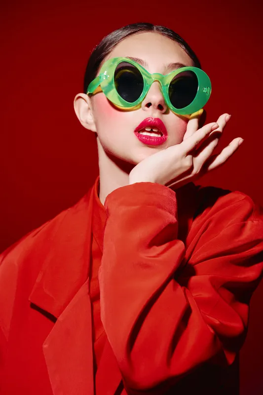 Elegant young woman wears vibrant green sunglasses and bold red lipstick. She poses in red outfit against red backdrop creating a strong beauty fashion creative concept.