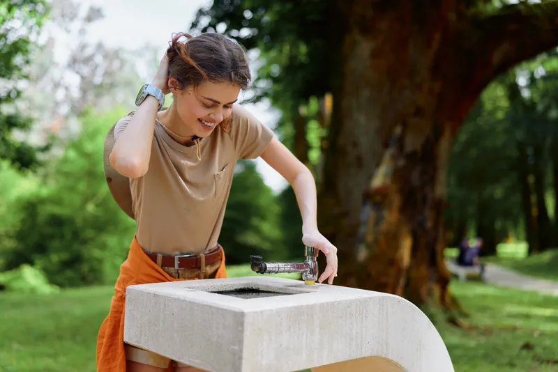 Young woman smiling and drinking from a public water fountain in a green park, casual summer outfit, pressing the spout for hydration, outdoor leisure and healthy lifestyle moment.
