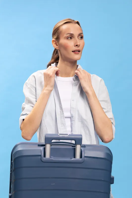 Young woman with blue suitcase looks away thoughtfully on blue background. Travel concept with casual clothes and preparation for journey or vacation with luggage.