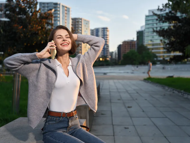 A cheerful woman talks on her phone outdoors in a city plaza, smiling with confidence as the modern skyline rises behind her, creating a bright, energetic urban moment.