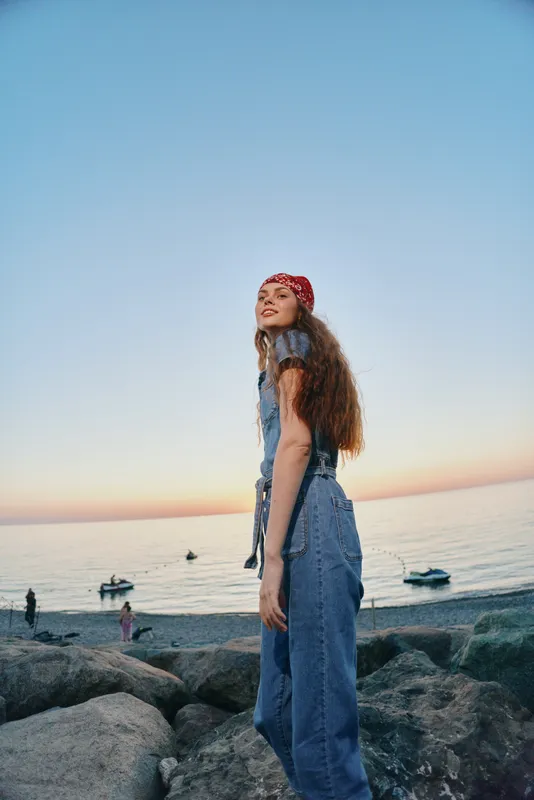 lifestyle woman in modern boho-western denim street style stands on rocky shore at sunset with film color tones and relaxed mood
