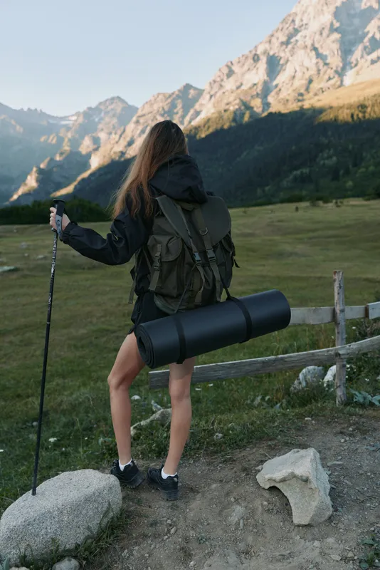 A lone hiker stands on a dirt trail in a rugged mountain landscape, with a trekking pole in hand and a rolled mat strapped to a sturdy backpack, ready for adventure and exploration.