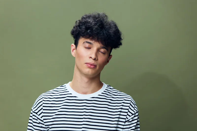 Young man with closed eyes wearing a striped shirt stands against a plain green background, expressing calmness and relaxation in a minimalistic setting.