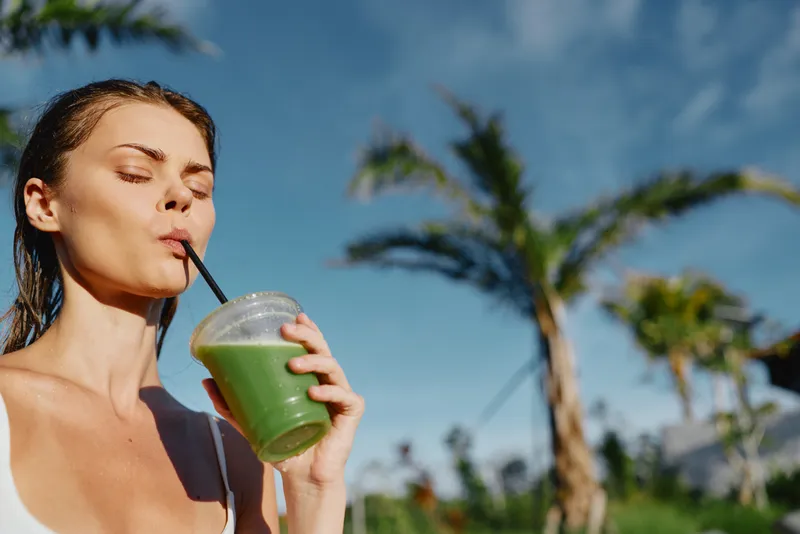A woman in a bright outdoor setting sips a green smoothie through a straw, enjoying a refreshing moment. A lifestyle scene focused on health, hydration, and sunny leisure.