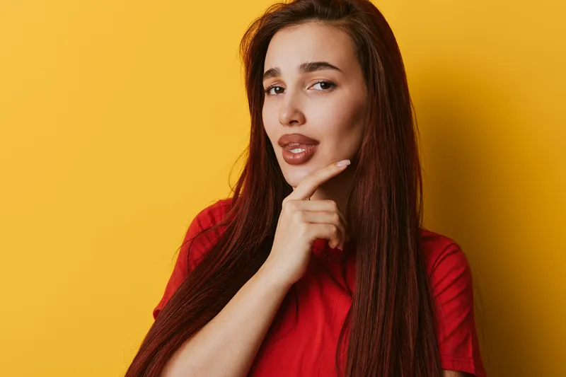 Woman with long hair and red shirt posing with thoughtful expression against yellow background. Female model looking at camera with hand on chin in studio portrait.
