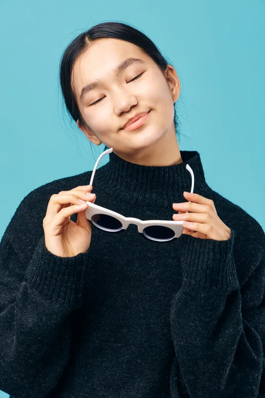 Young woman with closed eyes holding white sunglasses smiling with relaxed expression. Female person in black sweater enjoying moment with casual style and serenity on blue background.