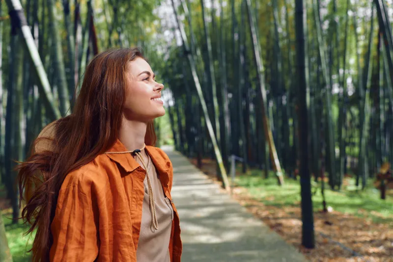 Woman with a smile and redhead hair wearing an orange jacket walking along a bamboo forest path, enjoying nature and sunlight in a peaceful outdoor portrait.