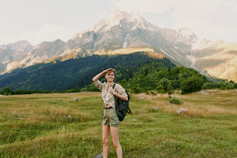 Young woman with a backpack explores a grassy valley, shielding eyes from sunlight as towering mountains rise behind her, enjoying a carefree outdoor moment.