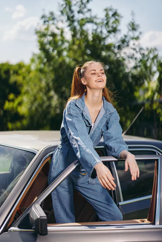 Young woman in denim jumpsuit joyfully leaning out of a vintage car against a sunny backdrop of greenery, embodying carefree summer vibes and happiness.