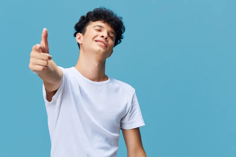 smiling young man pointing at camera in white t-shirt against blue background, happy expression, casual style, studio portrait