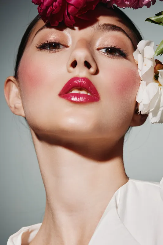 Close-up portrait of a young woman with glossy red lips and bronze eyeshadow, surrounded by vibrant pink and white flowers. Concept of creative beauty and fashion artistry.
