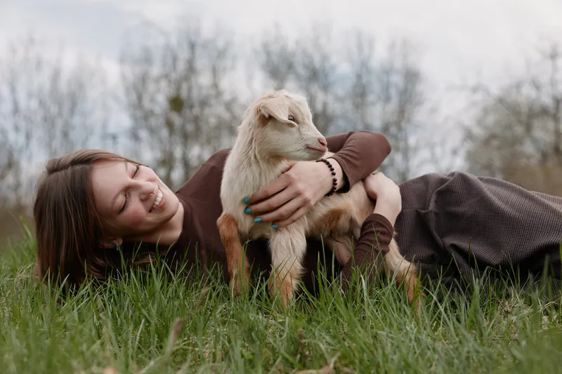 Smiling woman lying in grass, cuddling a goat kid in a sunlit meadow. Casual lifestyle moment highlighting animal bond, nature, and gentle affection in an outdoor setting.