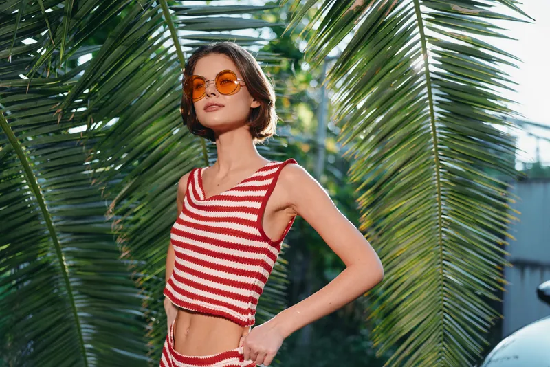 A stylish young woman stands outdoors among tropical palm leaves, wearing a red and white striped crop top and sunglasses. She poses confidently, capturing a vibrant, sunny, fashion-forward moment