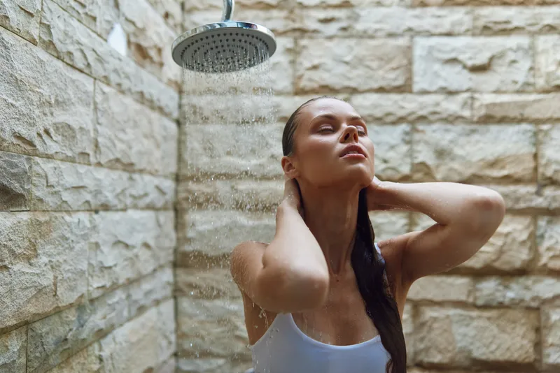 Young woman taking a relaxing shower under rainfall showerhead in modern stone bathroom, enjoying water droplets and serene moment with eyes closed and hands behind head.
