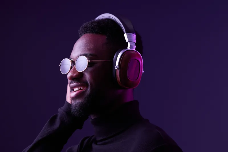 A smiling young Black man wearing trendy glasses and headphones, enjoying music against a vibrant purple background, conveying modern lifestyle and joy