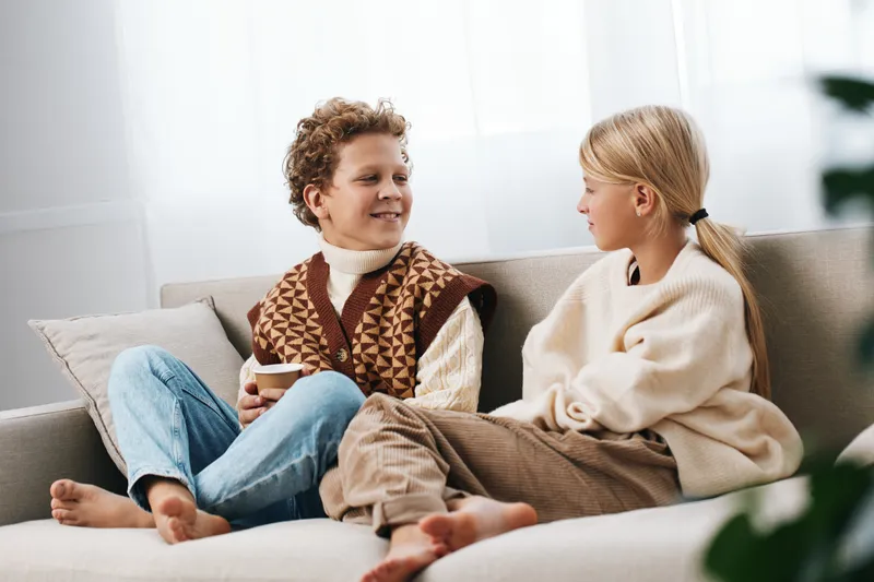 Two children share a cozy moment on a sofa, smiling and chatting while dressed in warm sweaters and jeans, creating a candid scene of comfort and friendship.