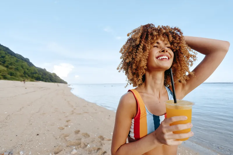 Smiling woman with curly hair on sunny beach, seaside background, holding a refreshing drink with straw, arms behind head in relaxed pose, summer vacation atmosphere, natural light