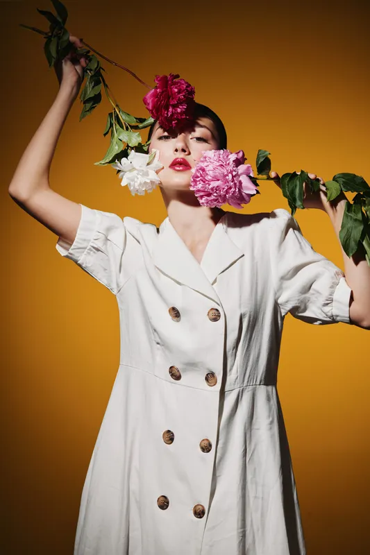 Young woman in a white dress holding vibrant peonies close to her face and their branches raised, posing studio fashion portrait on yellow background with creative beauty concept.
