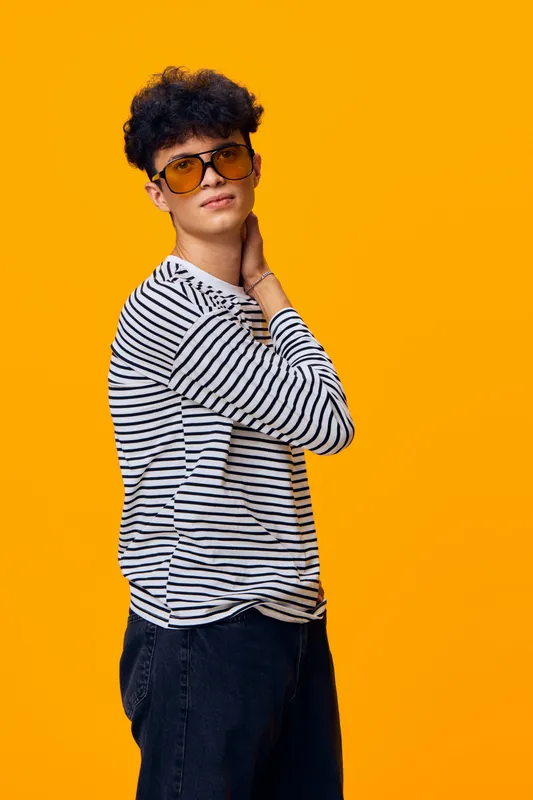 Young man in striped shirt wearing trendy sunglasses poses confidently against vibrant yellow background, showcasing casual style and modern fashion sense.