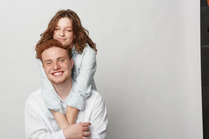 Happy young couple with red hair and casual clothes smiling and hugging each other on light background. Concept of love, friendship, relationship, and positive emotions between people.