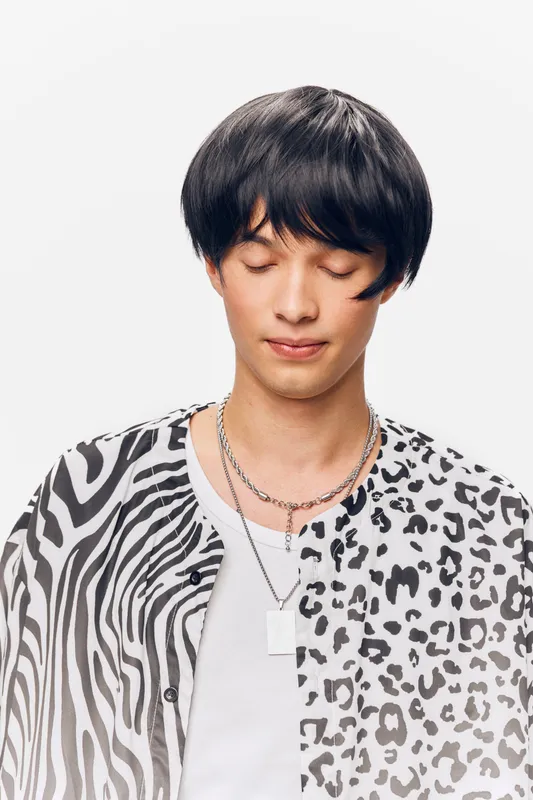 calm man with closed eyes wearing a stylish black and white animal print shirt and silver necklaces against a plain white background, expressing peacefulness and tranquility.