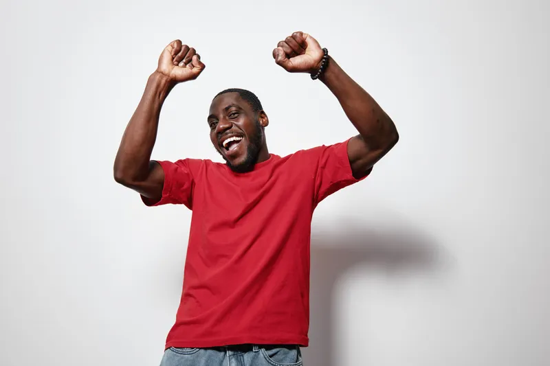 African American man with dark skin wearing red t shirt dancing and smiling with raised hands isolated on white background for happiness and celebration.