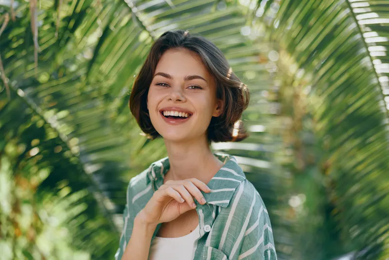 Young woman with short dark hair smiling brightly in a sunny tropical setting, wearing a striped green shirt over a white top.