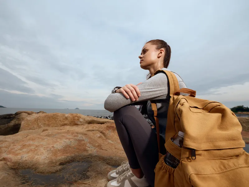 Thoughtful young woman sitting on rocks by the sea with a backpack, wearing casual clothing and capturing a serene outdoor moment