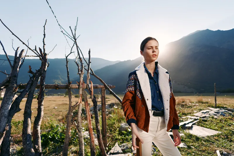 Woman standing outdoors with mountains and rustic fence, portrait with sun backlight, nature scenery and casual fashion outfit in an autumn field with contemplative expression.