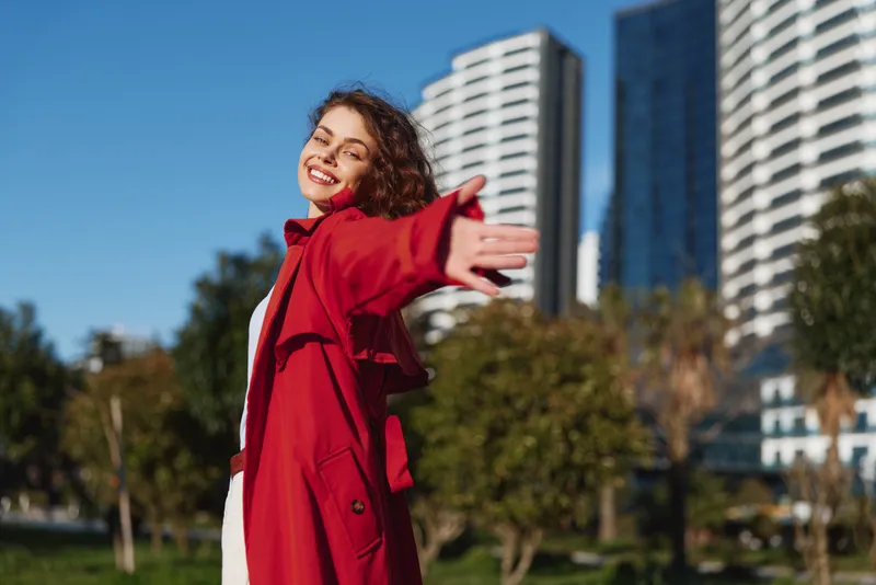 Young woman in a red coat smiles with open arms in an urban park, sunlight highlights her expression as modern buildings rise behind, conveying freedom.