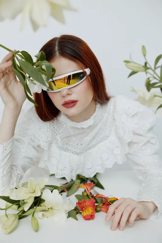 A stylish woman with bold red hair and retro sunglasses poses among fresh flowers, showcasing a blend of modern fashion and nature s beauty, ideal for seasonal campaigns