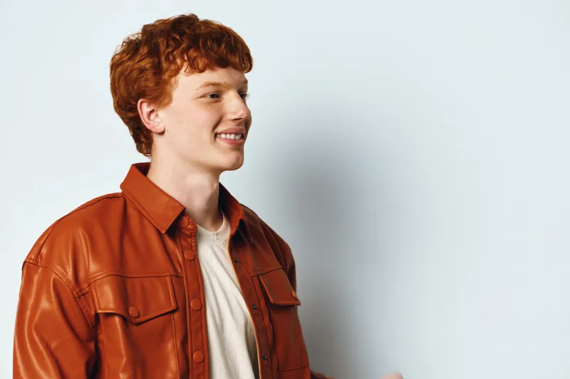 Young man with red hair wearing brown leather jacket and white shirt standing and smiling on light gray background. Casual style, positive expression and happy mood for portrait or fashion.
