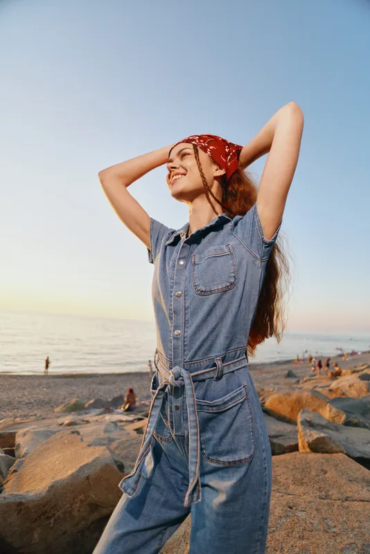 young woman smiling wearing red bandana and denim jumpsuit standing on rocky beach with ocean background during golden hour