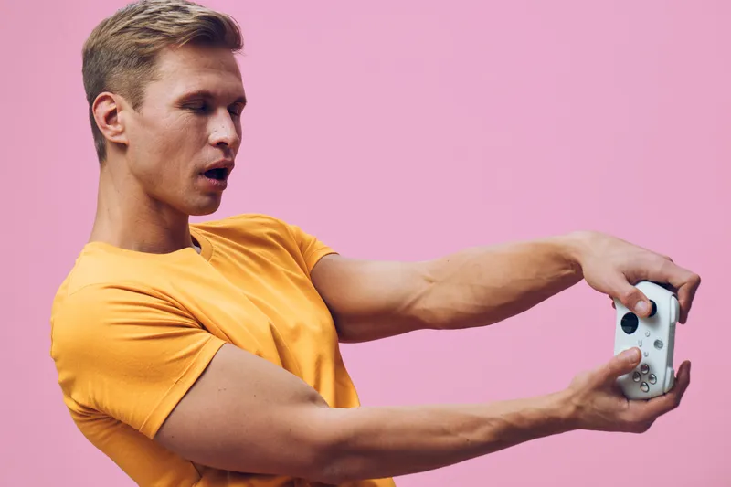 Young man with blond hair wearing orange t-shirt holding a gaming controller, standing against pink background, experiencing concentration during gameplay, studio portrait, lifestyle concept, vibrant 
