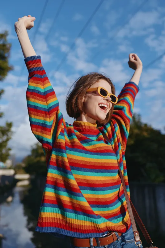 Young woman in a rainbow sweater and yellow sunglasses, smiling with arms raised outdoors by a canal; casual outfit, short hair, carefree travel and lifestyle moment under sunny blue sky.