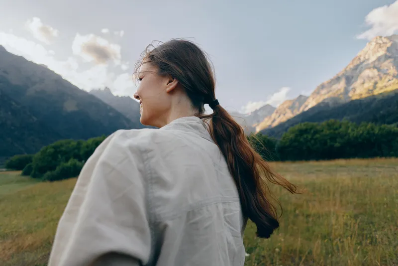 Woman in casual shirt stands in a meadow with mountains in the background, capturing a serene outdoors lifestyle moment in nature, hair tied back and bright sky